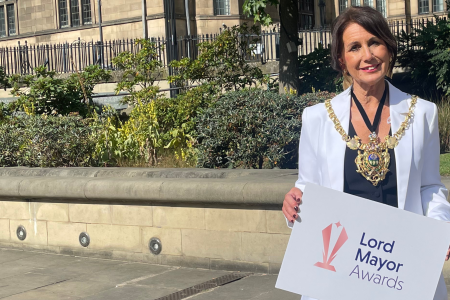 The Lord Mayor of Sheffield, Jayne Dunn, smiles at us while standing in the sunshone in the Peace Gardens. Dressed in a crisp white suit adorned by the gleaming golden mayoral chains she holds a sign with the Lord Mayor Awards logo on it.