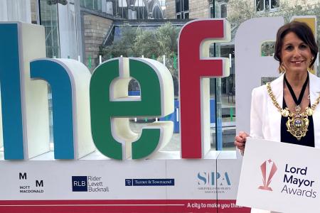 Lord Mayor of Sheffield, Jayne Dunn smiles in front of the multi-colour Sheffield sign in the Winter Gardens. She's holding a sign saying Lord Mayor Awards 
